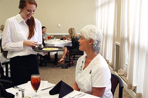 An attentive waitress serves food to a senior woman in a college dining setting.