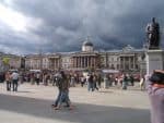 Historic building with large crowd and statue in foreground, under dark stormy clouds.