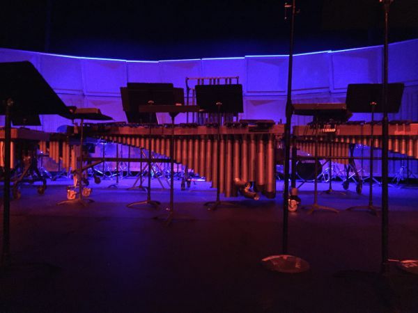 Percussion ensemble performing on stage with marimbas and drums illuminated by blue stage lighting.