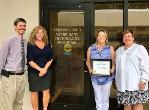 Group of four people standing outside Building 3200, Veterinary Technology Program, holding a certif.
