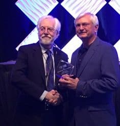 Two men in suits shaking hands at awards ceremony, celebrating welding program recognition.
