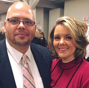 Bald man in glasses and suit with woman in pink at college event.
