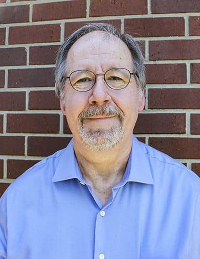 Music instructor with glasses and beard standing against brick wall, smiling.