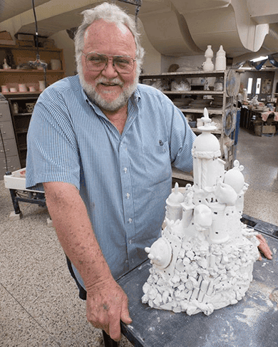 Senior man with white hair and beard, smiling, standing in ceramics studio with a white ceramic scul.