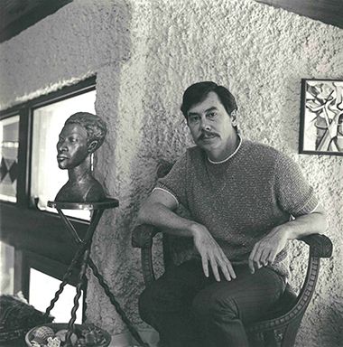 Portrait of a man sitting beside a bust sculpture in an art studio setting.