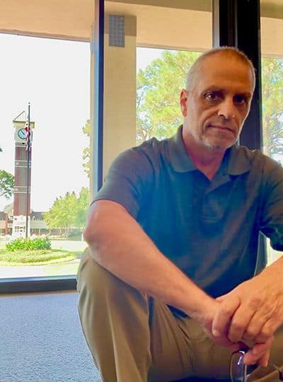 Accessible image of Troy Moon, an older man, sitting indoors with a window view of trees and a build.