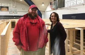 Two individuals standing inside a gymnasium with wooden bleachers and basketball hoops, supporting s.