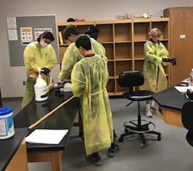 Students in yellow protective gowns practicing wound simulation with medical supplies and tools.
