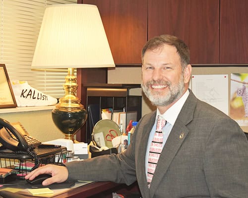 College administrator portrait in office with desk and lamp.