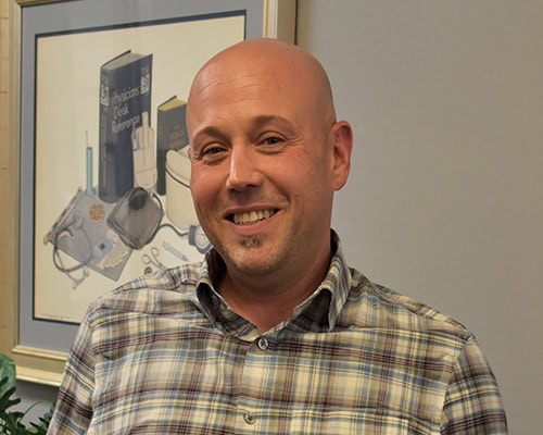Bald male college administrator smiling in office setting with framed educational artwork in backgro.