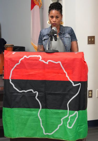 African American woman speaking at Black History Month event with African continent flag.