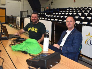 Two men seated at a table with audio equipment in a gymnasium setting.