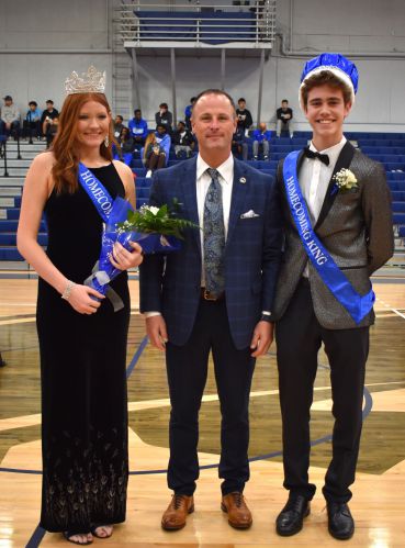 Prom King and Queen with school official during homecoming celebration in gymnasium.