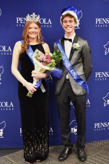 Smiling young man and woman in formal attire with crowns and sashes at college event.