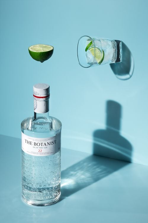 Lime wedge and gin bottle with glass of lime-infused water on blue background.