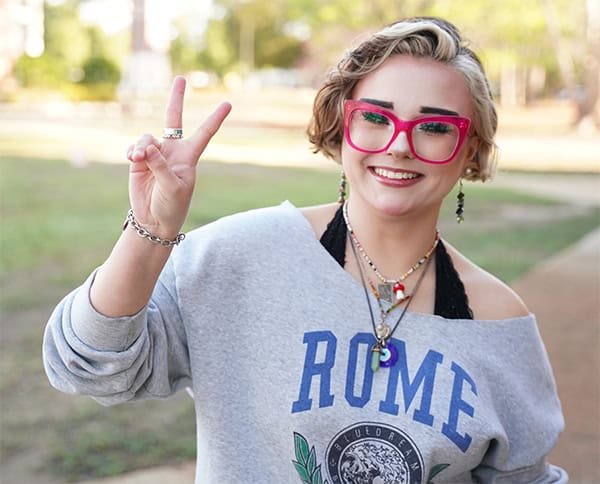 Student Jenny Woods smiling outdoors, making peace sign, showcasing college spirit and diversity.