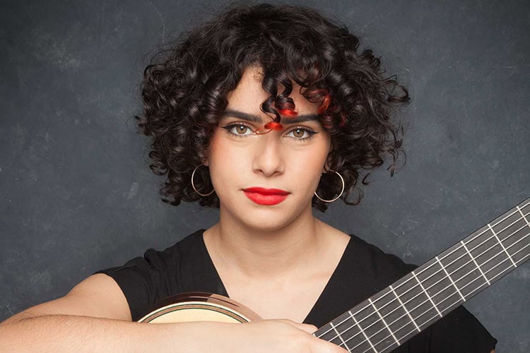 Female musician with curly hair playing acoustic guitar, promoting arts and music programs at Pensac.